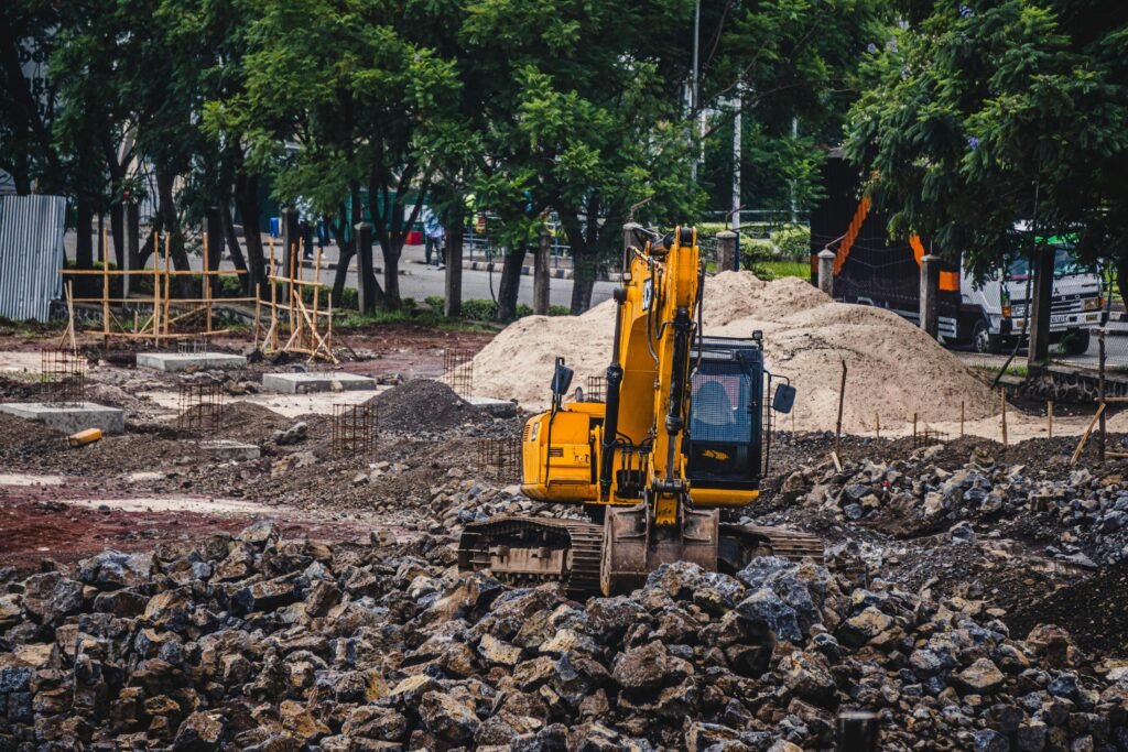 A yellow excavator working at an urban construction site surrounded by rocks and trees.